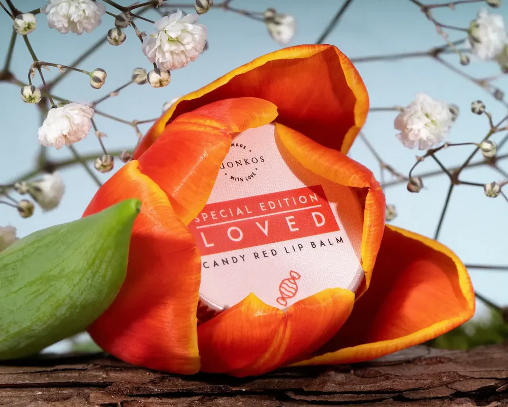 A red and orange tulip partially encircles a round tin of Luonkos Loved Special Edition Candy Red Lip Balm. White baby's breath flowers and a wooden surface appear in the background.