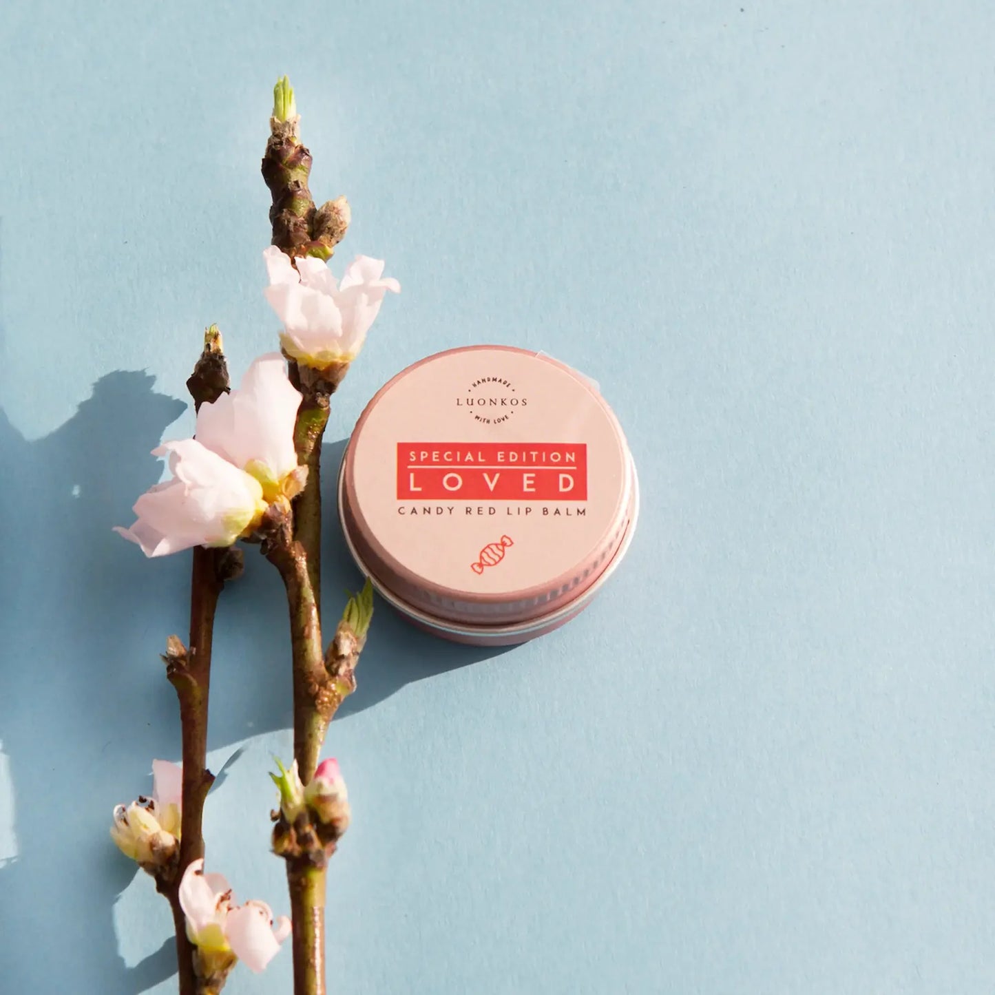 A tin of Luonkos Loved Special Edition Candy Red Lip Balm by Luonkos rests on a light blue surface beside a branch with pink blossoms.