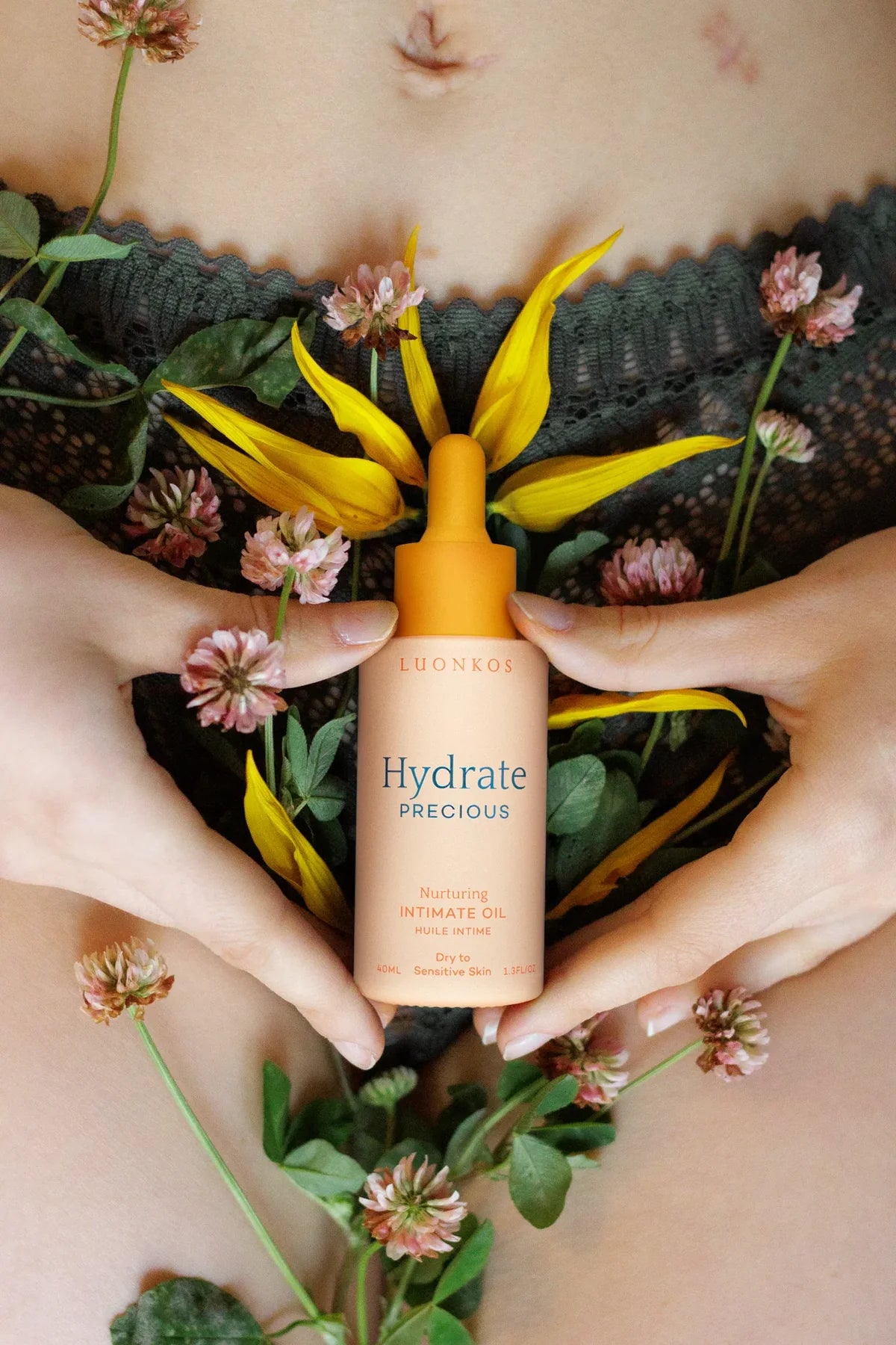 A person holds a bottle of Luonkos Precious Intimate Oil by Luonkos against their lower abdomen, surrounded by yellow petals and pink clover flowers, with green lace underwear visible—ideal for sensitive mucous membranes.