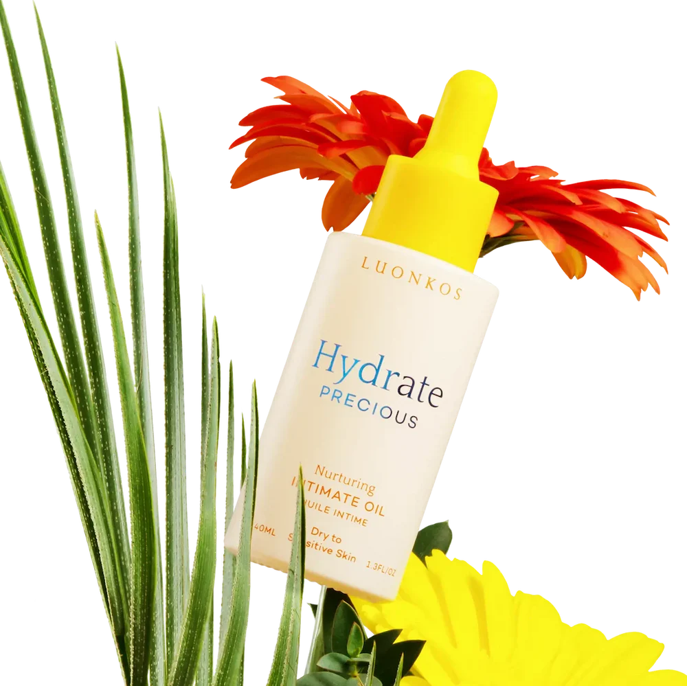 A bottle of Luonkos Precious Intimate Oil, ideal for sensitive skin and dryness, rests among green palm leaves and bright orange and yellow flowers on a white background.
