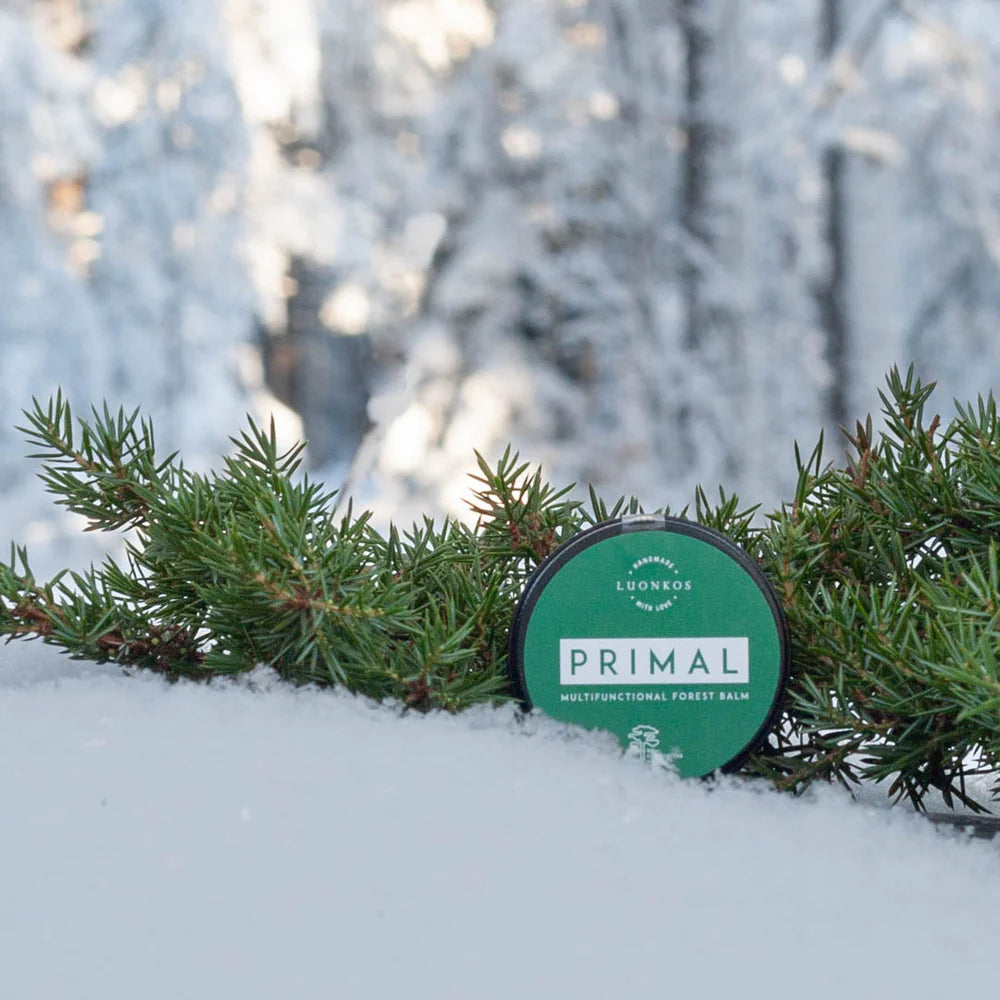 A round tin of Luonkos Primal Multifunctional Forest Balm rests on snowy ground among evergreen branches, emphasizing its natural ingredients for soothing eczema-prone skin, with a blurred snowy forest backdrop.