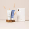 A baby gift set from MeiForest featuring a white and blue lotion/body wash tube set displayed on a wooden slice stand next to a white gift box with a small duck logo, decorated with dried pampas grass against a warm beige background.