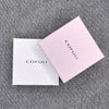 Two square boxes—one white and one light pink—both labeled "Coruu" in black, rest on gray fabric. The pink box slightly overlaps the white, displaying stylish packaging for Coruu Moomin Wooden Earrings.
