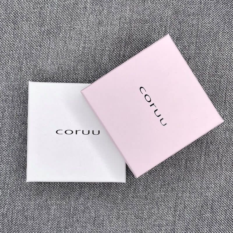 Two square boxes—one white and one light pink—both labeled "Coruu" in black, rest on gray fabric. The pink box slightly overlaps the white, displaying stylish packaging for Coruu Moomin Wooden Earrings.