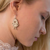 Close-up of a woman with light hair looking down, wearing Coruu Moomin Wooden Earrings shaped like a cartoonish monster from Coruu. The playful earrings are crafted from wood and feature a mischievous Moomin character.