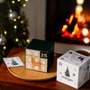 The Nordqvist Tea Advent Calendar sits on a tray beside a Nordqvist tea box, with a glowing Christmas tree and cozy fireplace in the background, creating a festive setting for the perfect Christmas tea gift.