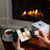 A person holds the Nordqvist Tea Advent Calendar with numbered drawers in front of a cozy fireplace, while a cup of Nordqvist tea sits on the table and another hand lifts drawer 5, adding festive cheer.