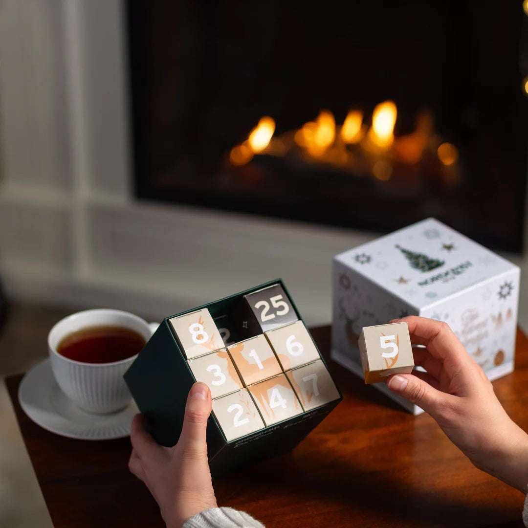 A person holds the Nordqvist Tea Advent Calendar with numbered drawers in front of a cozy fireplace, while a cup of Nordqvist tea sits on the table and another hand lifts drawer 5, adding festive cheer.