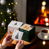 A person opens a Nordqvist Tea Advent Calendar decorated for Christmas beside a cup of Nordqvist tea on a wooden table, with a glowing fireplace and festive Christmas tree in the background.