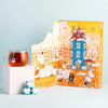 A glass mug of winter tea rests on a white cube next to the Nordqvist Moomin Advent Tea Calendar, showing Moomin characters in a snowy scene. Three small blue heart-patterned boxes add a cozy touch to the vibrant Nordqvist display.