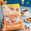 A person holds the Nordqvist Moomin Advent Tea Calendar, a colorful vegan tea selection by Nordqvist, on a blue table with a cup of tea, pastry, and a blue heart-shaped origami. Warm yellow lights glow in the background.