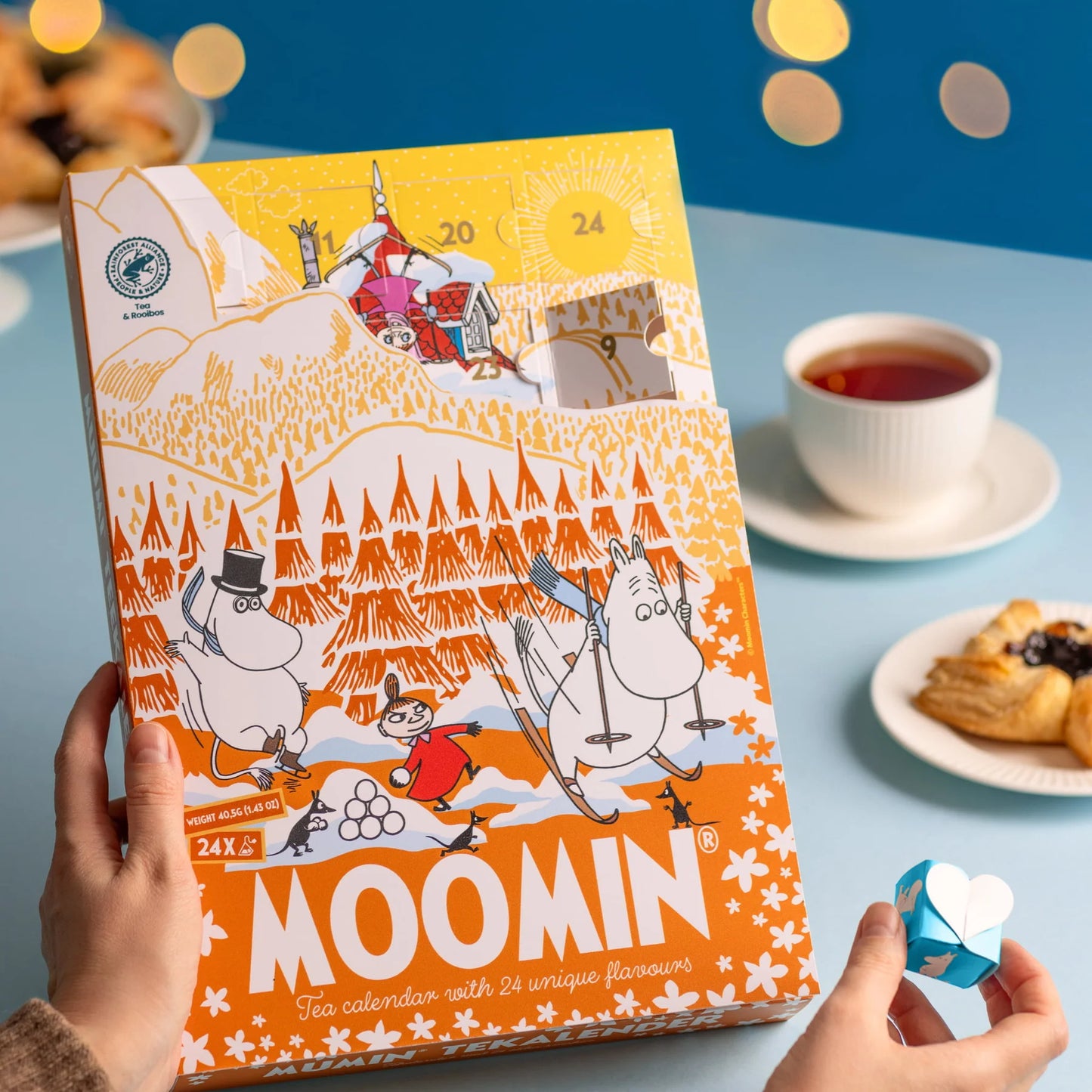 A person holds the Nordqvist Moomin Advent Tea Calendar, a colorful vegan tea selection by Nordqvist, on a blue table with a cup of tea, pastry, and a blue heart-shaped origami. Warm yellow lights glow in the background.