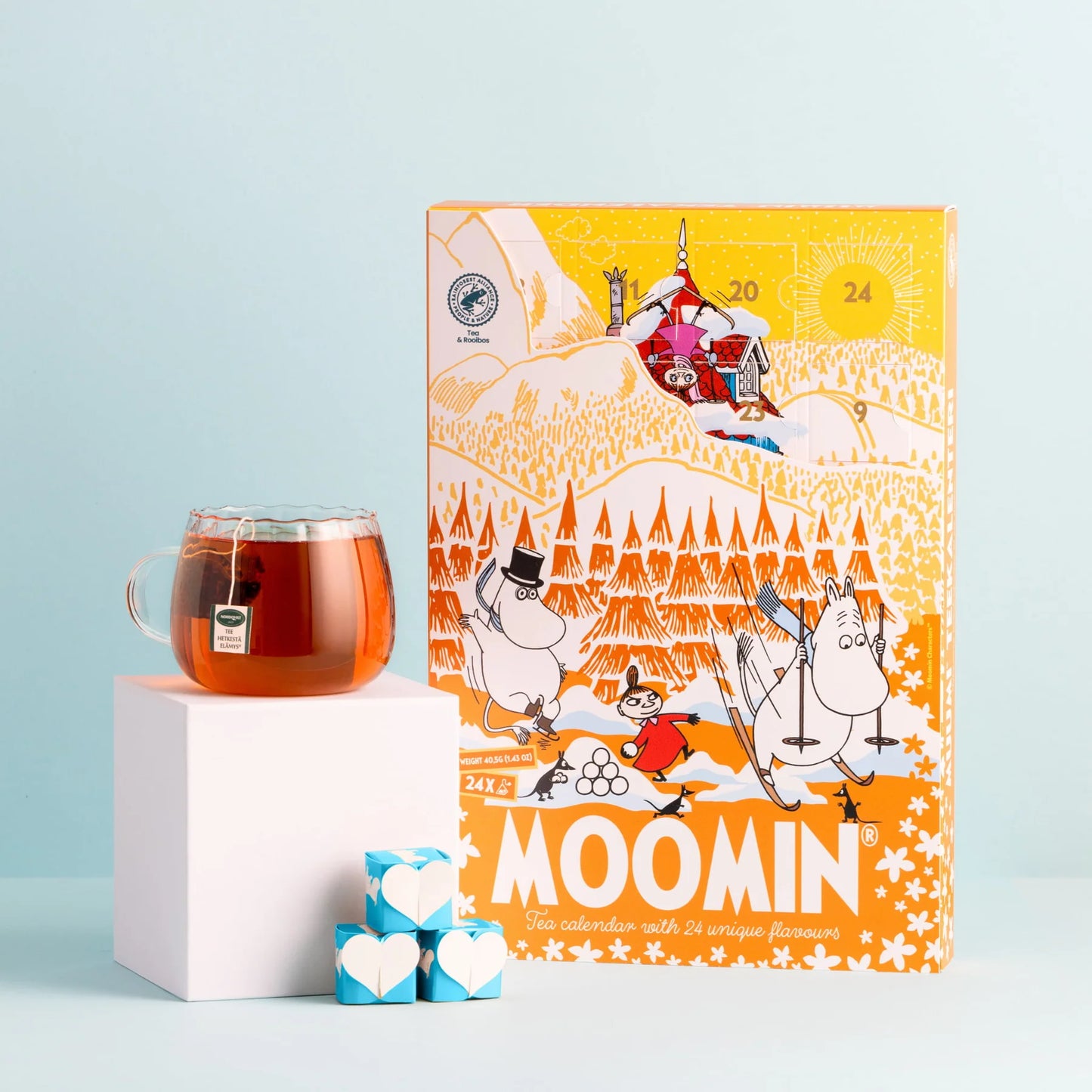 A glass mug of tea rests on a white block beside the Nordqvist Moomin Advent Tea Calendar and three blue boxes with white hearts, all set against a light blue background—ideal for enjoying a cozy vegan tea selection from Nordqvist.