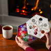 A person holds an open Nordqvist Winter Tea Collection with colorful, snowflake-patterned packaging next to a cup of tea on a wooden table by a lit fireplace.