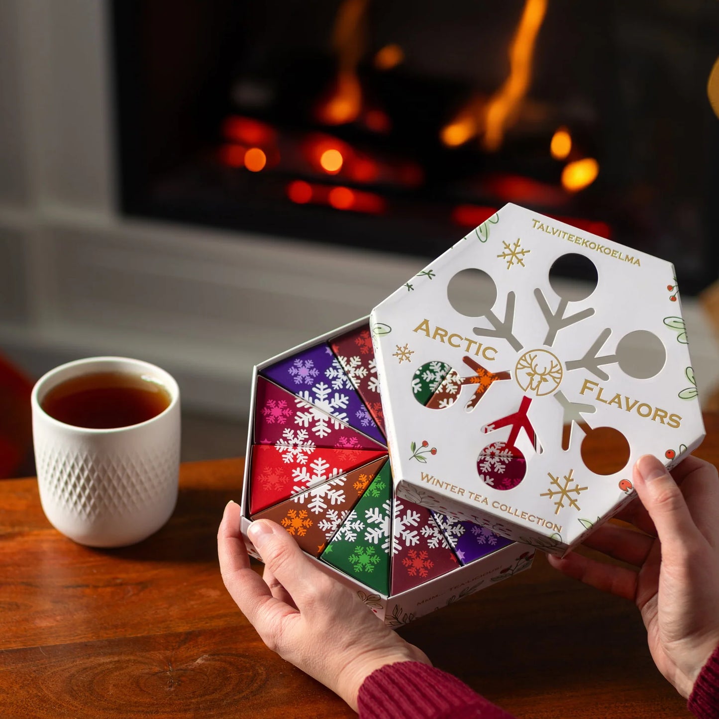 A person holds an open Nordqvist Winter Tea Collection with colorful, snowflake-patterned packaging next to a cup of tea on a wooden table by a lit fireplace.