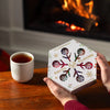 A person holds a hexagonal Nordqvist Winter Tea Collection box—an ideal holiday tea gift—near a wooden table with a white mug, with a fireplace and cozy atmosphere in the background.