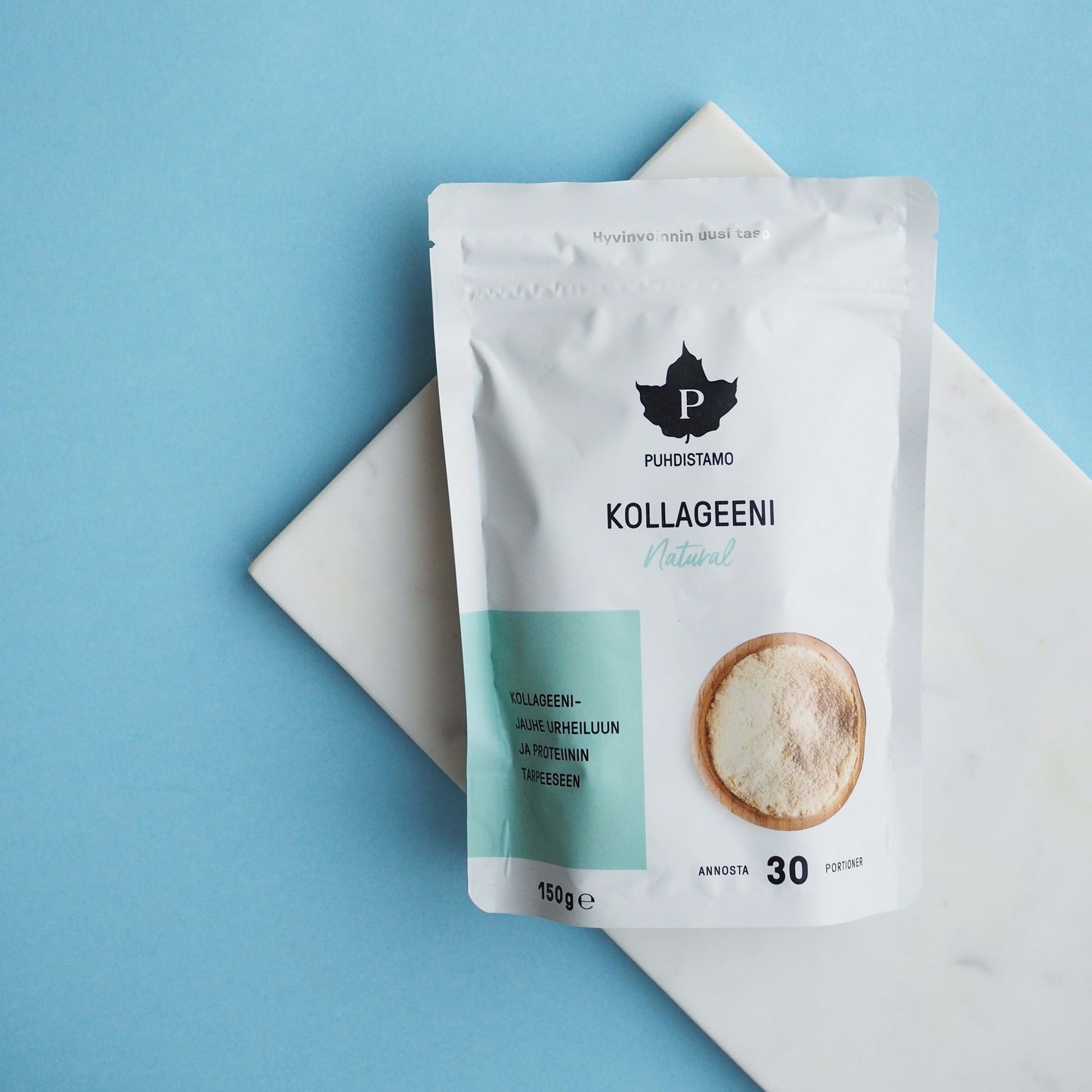 A white pouch of Puhdistamo Collagen Natural by Puhdistamo rests on a white marble slab with a light blue background. The packaging displays a black leaf logo, Finnish text, and emphasizes its Bodybalance collagen peptide content.