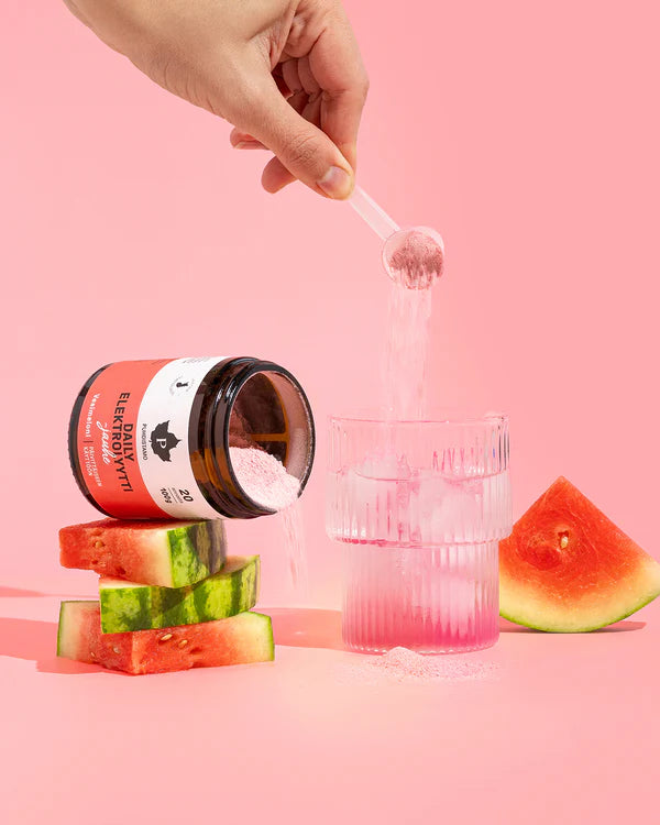 A hand pours Puhdistamo Daily Electrolyte Powder Watermelon into water beside an open jar labeled Puhdistamo, set on watermelon slices, highlighting daily hydration against a pink background.
