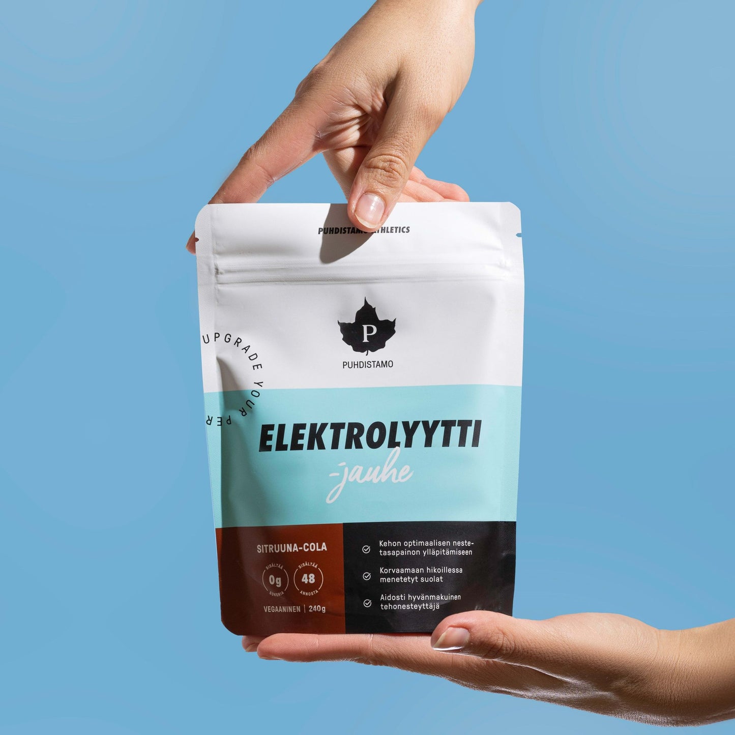 Hands hold a Puhdistamo Electrolyte Powder Lemon & Cola pouch labeled “ELEKTROLYYTTI jauhe” against a light blue background. The white and blue keto-friendly pack shows the Puhdistamo logo and hydration details.