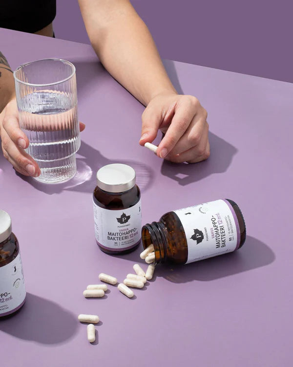 A person holding a glass of water gets ready to take a white capsule of Puhdistamo Strong Probiotic 12 Billion CFU, with several Puhdistamo probiotic bottles and spilled capsules on a purple surface, emphasizing gut health.