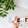 A hand holds white capsules of Puhdistamo Strong Probiotic 30 Billion CFU, poured from a Puhdistamo bottle over a marble countertop beside a glass of water and a leafy green plant.