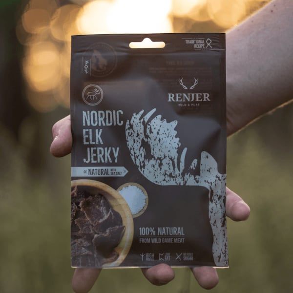 A hand holds a Renjer Nordic Elk Jerky Sea Salt package outdoors in warm sunlight. The brown packaging displays the Renjer brand, white text, and an image of elk jerky.