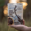 A hand holds a Renjer Nordic Reindeer Jerky Sea Salt pack outdoors, featuring reindeer antlers, "100% natural" text, and an image of sliced reindeer meat in a bowl.
