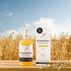 A bottle and box of Rimita Green’s RimitaOats Hand and Body Soap sit on a wooden surface outdoors, backed by a golden oat field and blue sky. The packaging displays Finnish oats illustrations and gentle cleansing claims for sensitive skin.