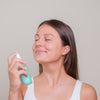 A woman with long brown hair and a white tank top sprays Rimita Green’s RimitaOats Facial Mist for deep hydration onto her face, eyes closed and smiling, against a plain light background.