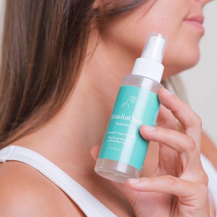 A woman in a white tank top holds a Rimita Green RimitaOats Facial Mist bottle with an aqua green label and white text near her face, about to spray the natural vegan mist with hyaluronic acid for deep hydration.