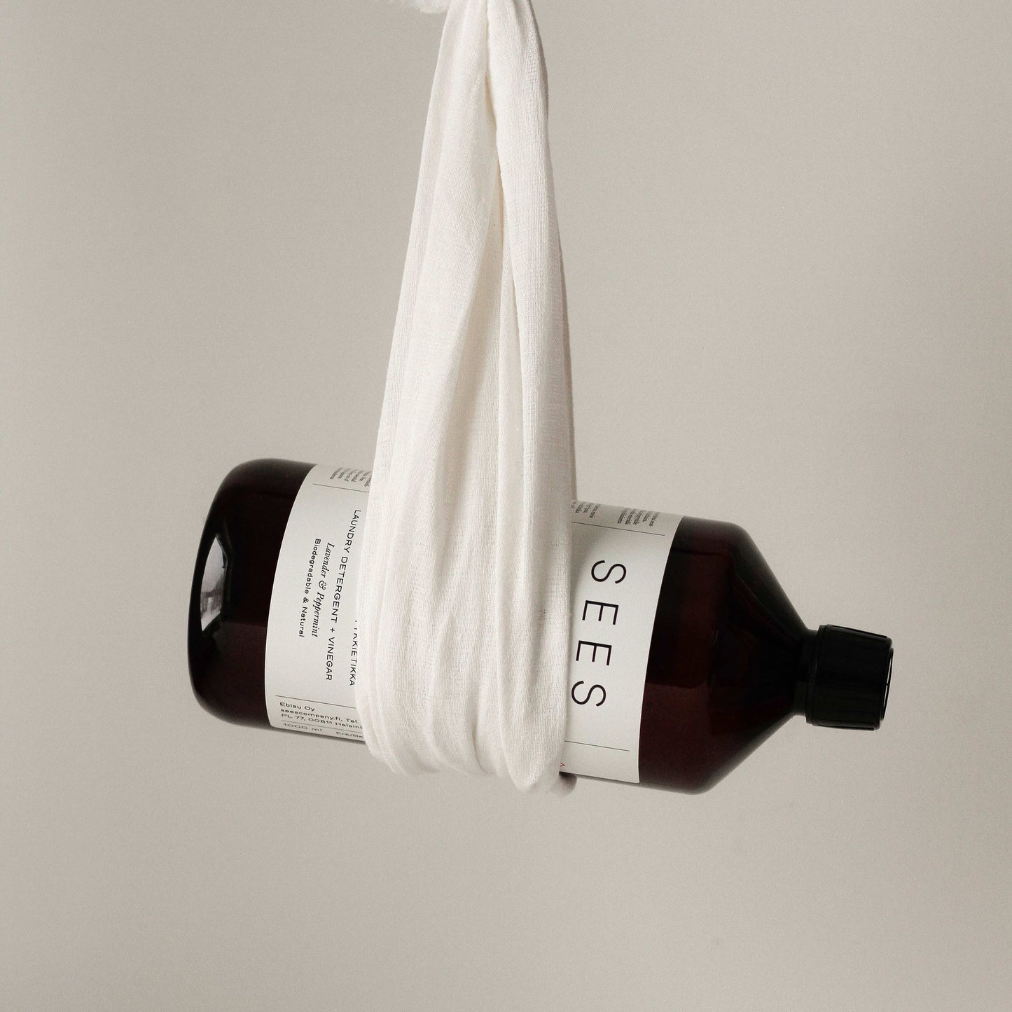 A dark brown SEES Company bottle labeled "SEES 2-in-1 Laundry Detergent with Vinegar" is wrapped in a white cloth and suspended against a plain, light background.