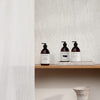 Three amber pump bottles of SEES Company’s SEES Body Wash, Cedar & Orange, with minimalist labels, are arranged on a wooden shelf against a textured white wall beside a white curtain and a pale ceramic vase below the shelf.