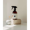 A brown spray bottle of SEES Company’s plant-based Cleaning Spray for the Kitchen and Bathroom sits on a stone block next to a small white vase with dried flowers, softly lit against a pale wall.