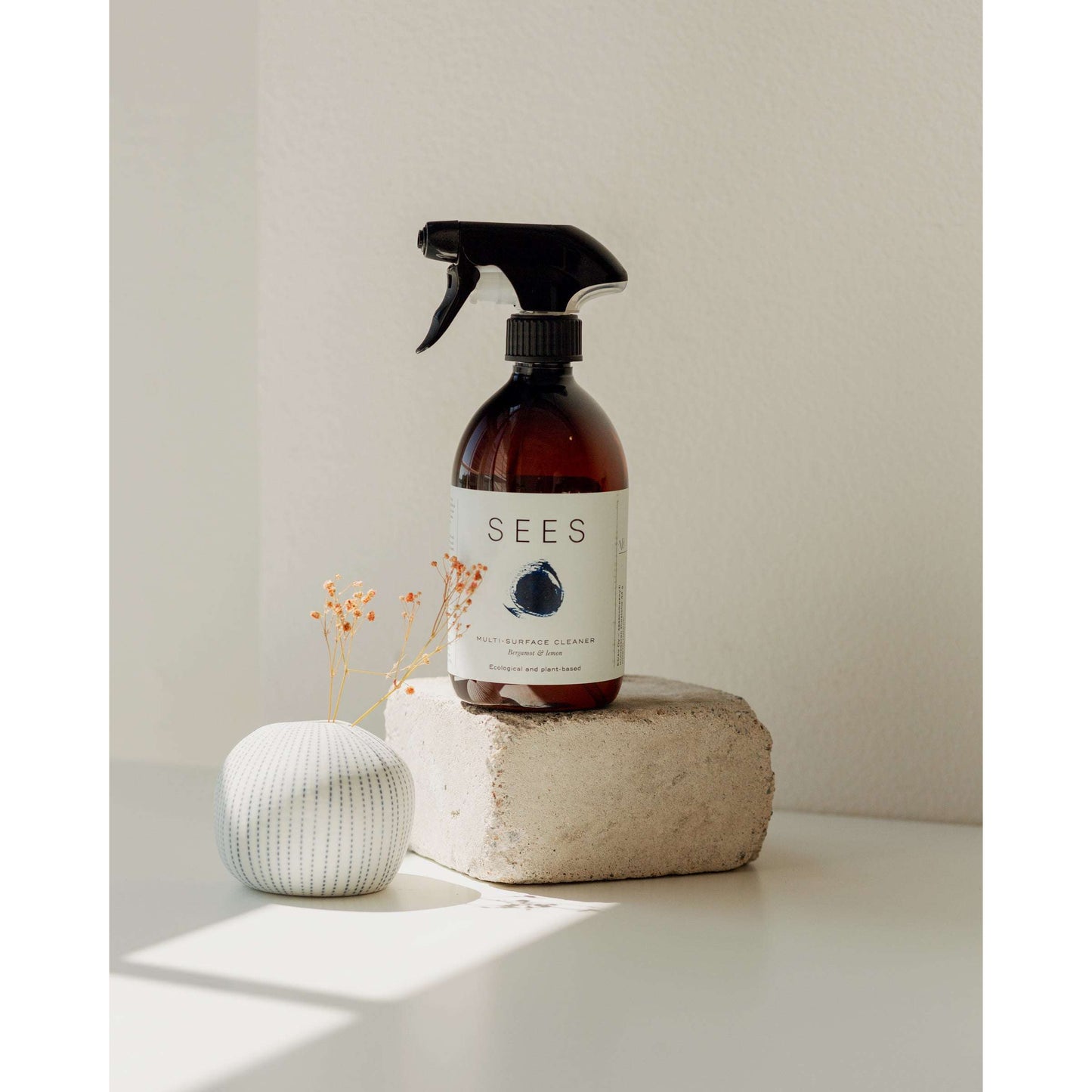 A brown spray bottle of SEES Company’s plant-based Cleaning Spray for the Kitchen and Bathroom sits on a stone block next to a small white vase with dried flowers, softly lit against a pale wall.