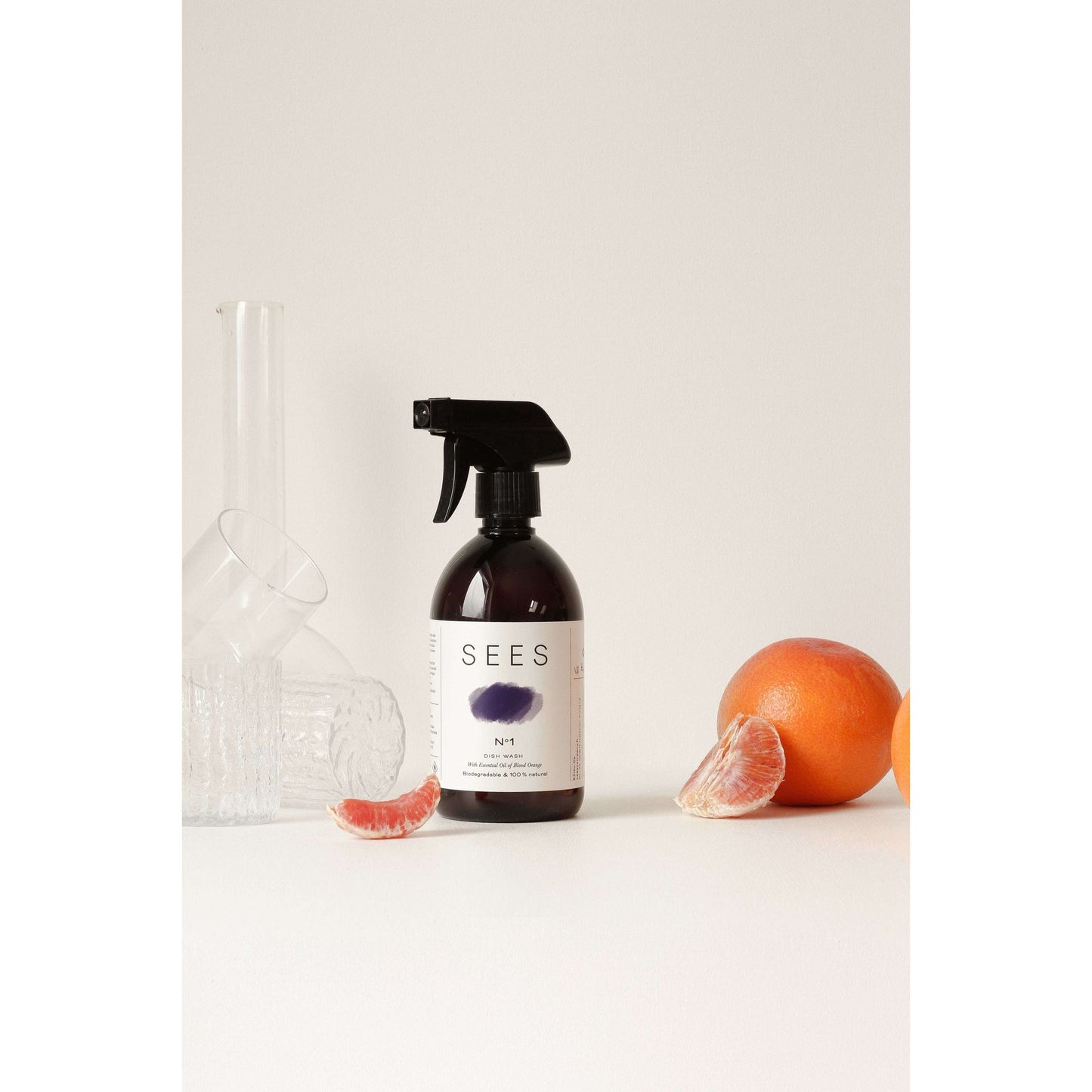 A SEES Company spray bottle of SEES Dishwashing Liquid sits on a white surface with glassware and grapefruit, emphasizing its eco-friendly, biodegradable cleaning amidst a simple light backdrop.