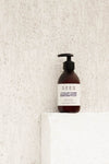 A dark brown pump bottle labeled “SEES” with white and blue text sits on a white textured ledge against a white wall. The label reads “SEES Hand & Body Lotion” by SEES Company, featuring Lavender & Orange for natural, vegan skincare.