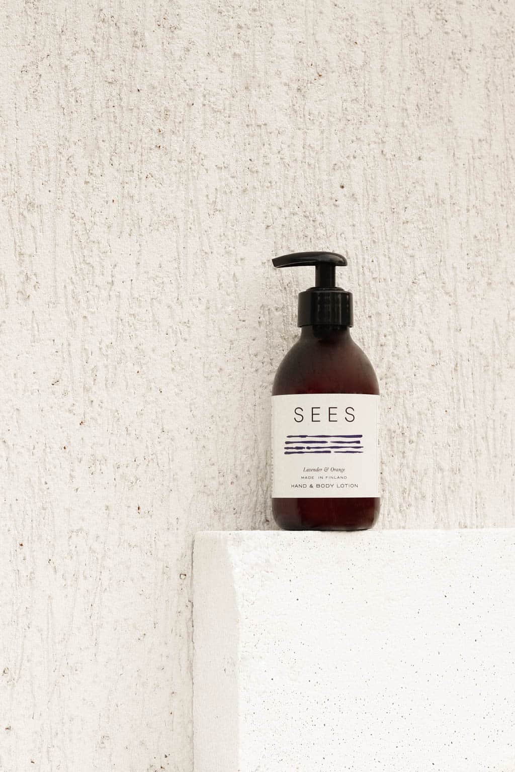 A dark brown pump bottle labeled “SEES” with white and blue text sits on a white textured ledge against a white wall. The label reads “SEES Hand & Body Lotion” by SEES Company, featuring Lavender & Orange for natural, vegan skincare.