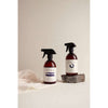 Two brown SEES Company spray bottles with white labels reading “SEES” sit on a light surface. One is SEES Textile Spray No. 1 Serene, Bergamot & Lemon, placed atop a small stone with sheer white cloth nearby. The background is plain and neutral.