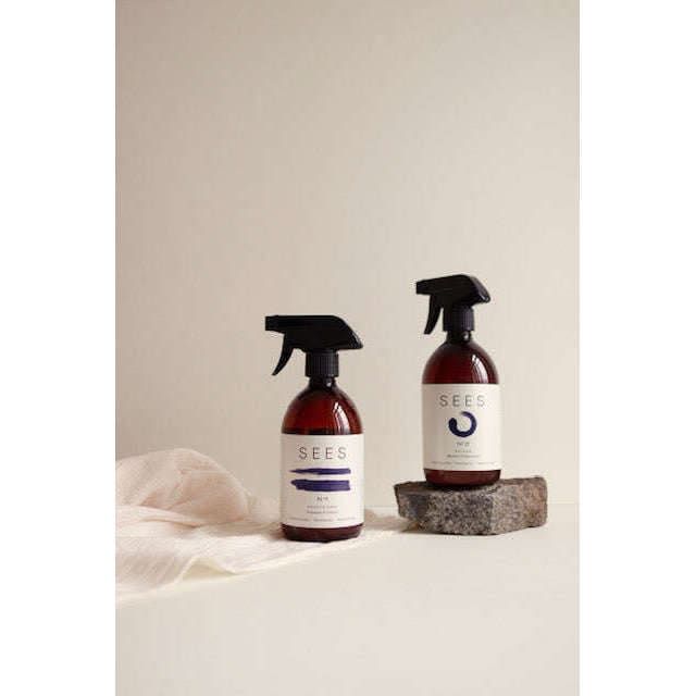 Two brown SEES Company spray bottles with white labels reading “SEES” sit on a light surface. One is SEES Textile Spray No. 1 Serene, Bergamot & Lemon, placed atop a small stone with sheer white cloth nearby. The background is plain and neutral.