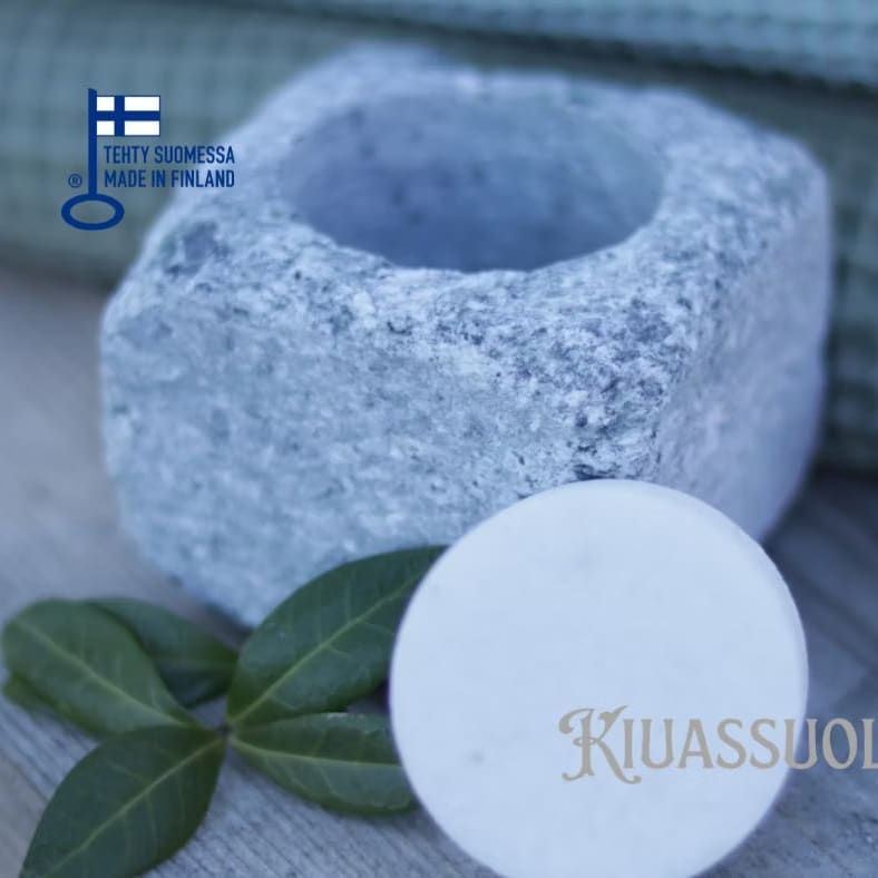 The Kiuassuola Sauna Salt ILMARI Starter Pack is displayed in a stone bowl with a white disk on wood beside green leaves. A green towel with Finnish and English text, plus a Finland flag and "KIUASSUOLA," appears in the background.