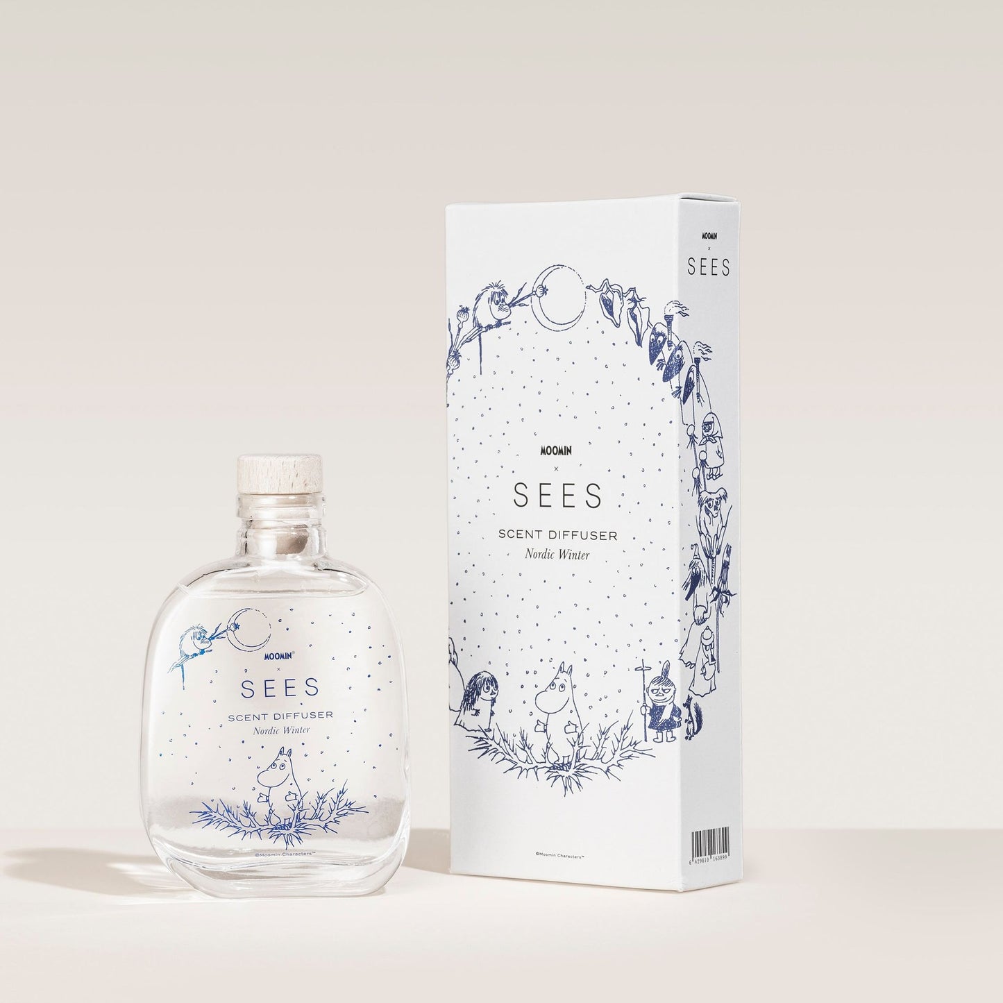 A clear glass SEES x Moomin Room Diffuser Nordic Winter by SEES Company, with a wooden cap, sits next to a white box featuring playful blue illustrations—a tribute to Finnish design and the cozy ambiance of Nordic winters.