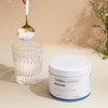A hand holds a spoonful of THERA Nordic Rezcue Drink Powder above a glass of water next to its white container on a beige surface, with delicate flowers in the background—ideal for gut healing and acid reflux relief.