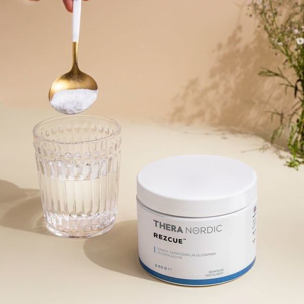 A hand holds a spoonful of THERA Nordic Rezcue Drink Powder above a glass of water next to its white container on a beige surface, with delicate flowers in the background—ideal for gut healing and acid reflux relief.