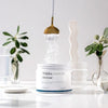 A hand pours THERA Nordic Rezcue Drink Powder from a spoon into an open container. The scene is bright, with glassware and green plants in the background, highlighting this gut-support product from THERA Nordic.