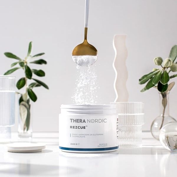 A hand pours THERA Nordic Rezcue Drink Powder from a spoon into an open container. The scene is bright, with glassware and green plants in the background, highlighting this gut-support product from THERA Nordic.