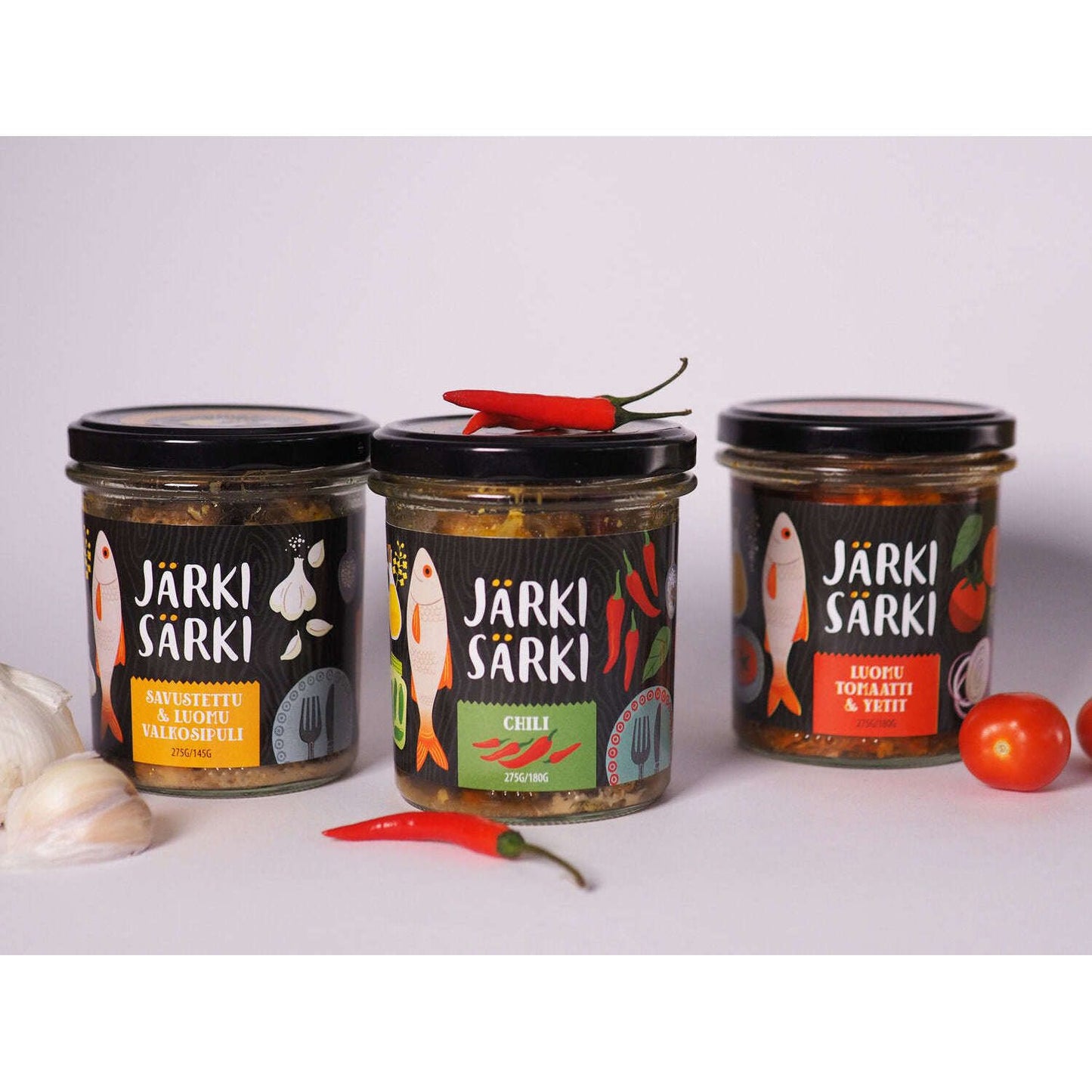 Three jars of Järki Särki Organic Tomatoes and Herbs preserves are displayed with garlic, chili peppers, and a tomato. The labels feature colorful fish illustrations highlighting the organic tomatoes and herbs flavor.