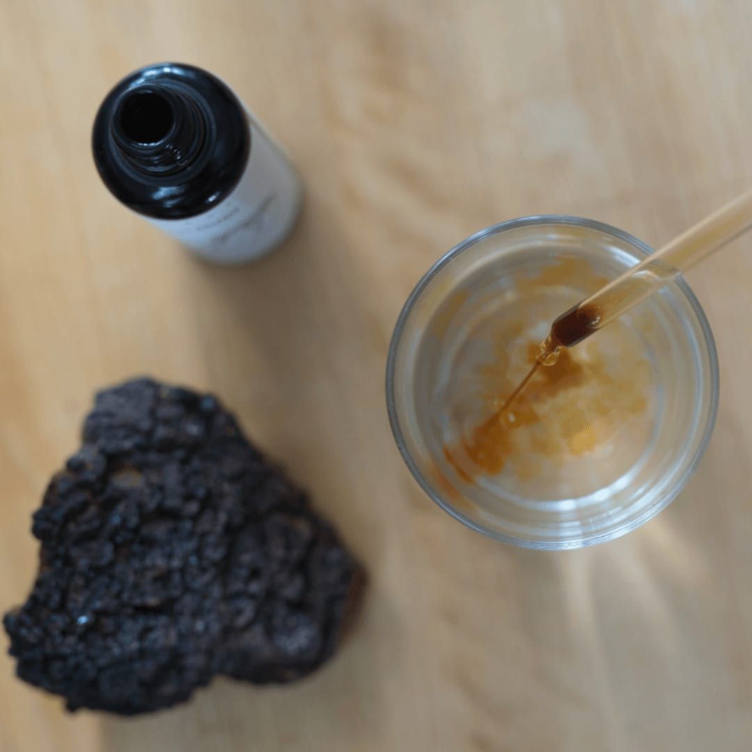 A pipette with amber Organic Chaga Extract from KÄÄPÄ Mushrooms hovers over a glass of water. Nearby, Finnish chaga and the extract’s open bottle rest on a light wooden surface.
