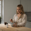 A woman sits at a minimalist kitchen table, stirring a drink with a dropper filled with KÄÄPÄ Mushrooms Organic Chaga Extract. A bottle of KÄÄPÄ Mushrooms Chaga and a glass of dark liquid are also on the softly lit table.