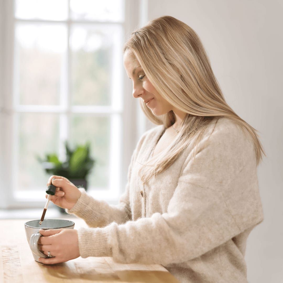 A woman with long blonde hair smiles as she adds KÄÄPÄ Mushrooms Organic Lion's Mane Extract to her mug at a wooden table, intent on brain health. A large window and potted plant create a softly lit, peaceful atmosphere.