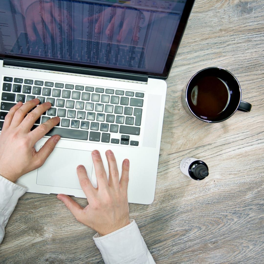 A person works on a laptop at a wooden desk with coffee and a small bottle of KÄÄPÄ Mushrooms Organic Lion's Mane Extract nearby, suggesting a focus on productivity and brain health, while the screen displays a detailed spreadsheet.